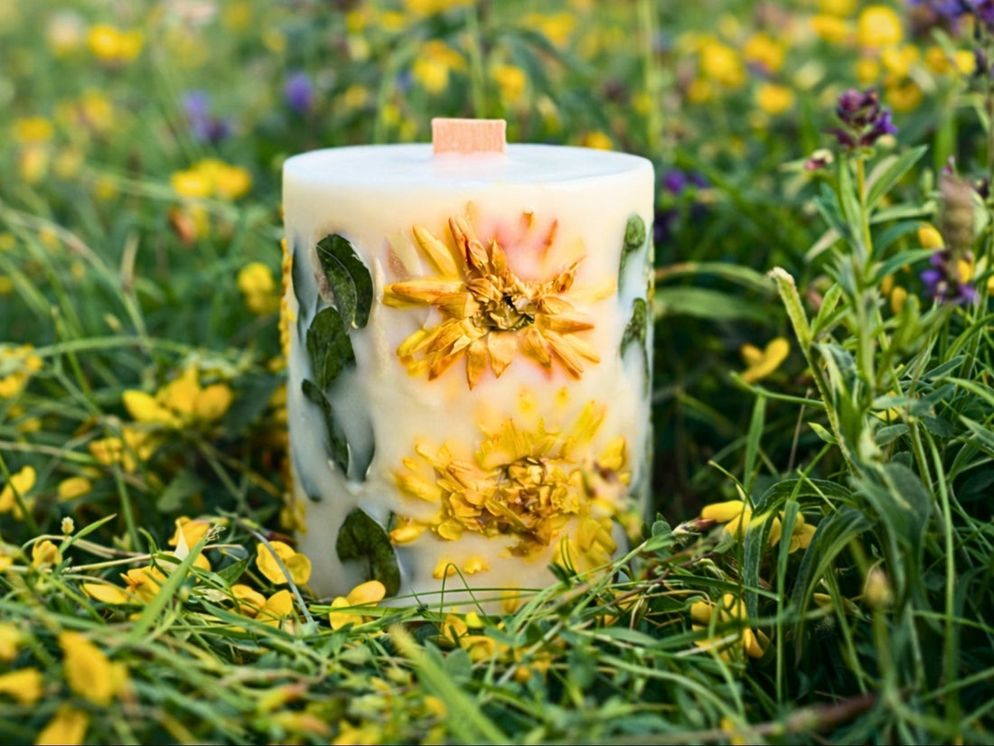 Handcrafted botanical pillar candle with real dried yellow flowers and green foliage embedded in natural soy wax, placed among wildflowers in a summer meadow. Hand-poured by Blossoms of Eden in Surrey.