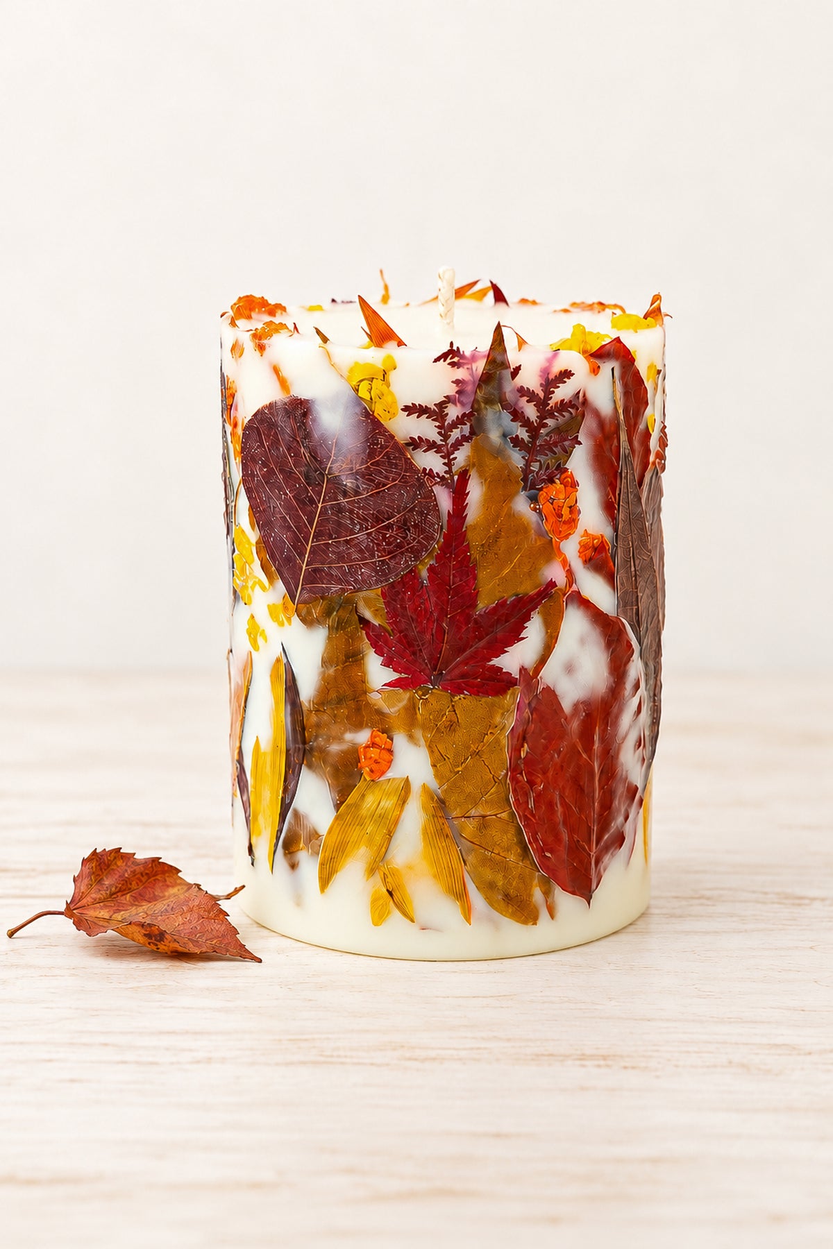 Botanical soy wax pillar candle decorated with real dried autumn leaves in red, orange, yellow and brown, standing on a pale wooden surface beside a fallen leaf.