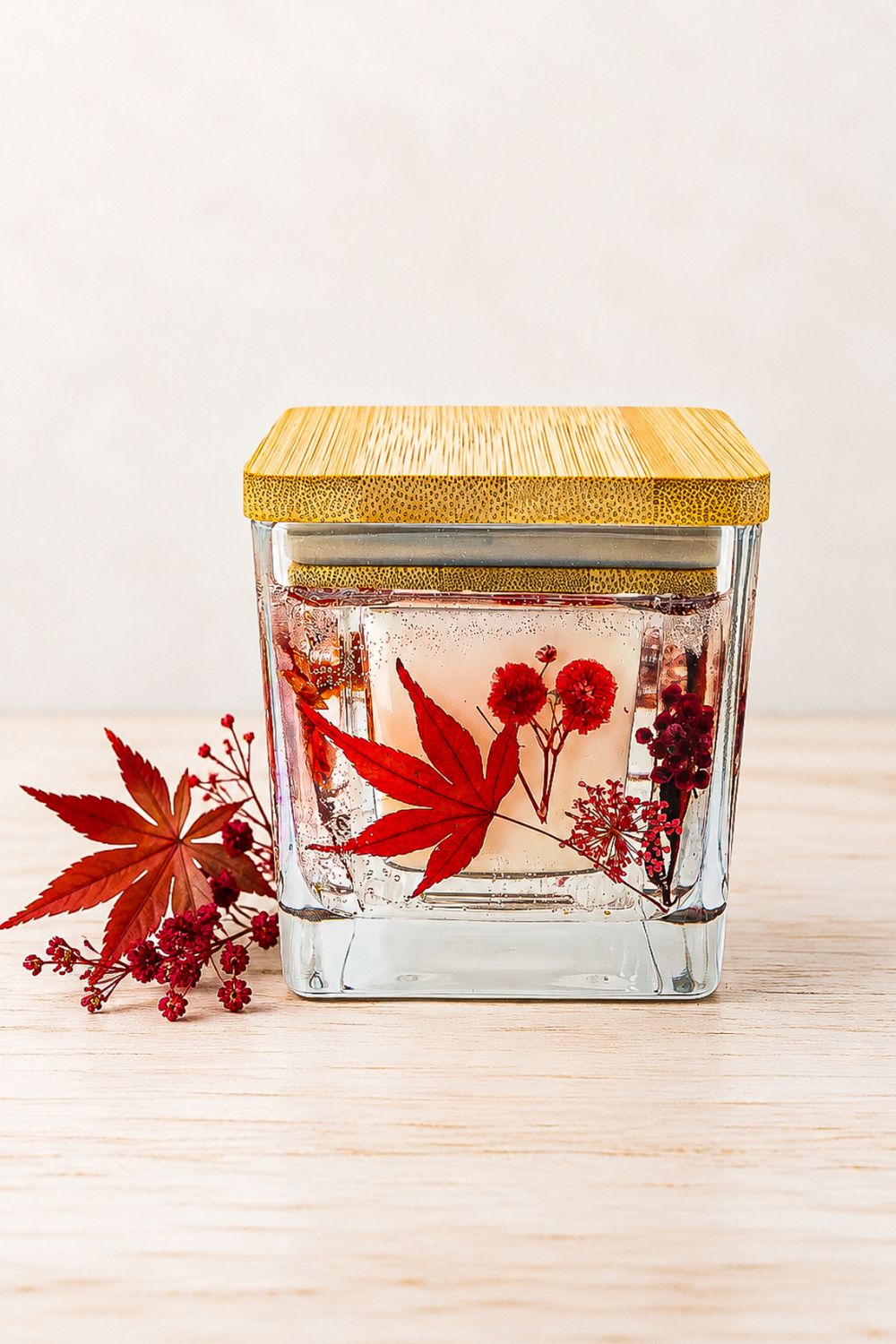 Autumn botanical soy wax jar candle with a red maple leaf and dried red flowers in a square glass container with a wooden lid, displayed on pale wood beside matching botanicals.