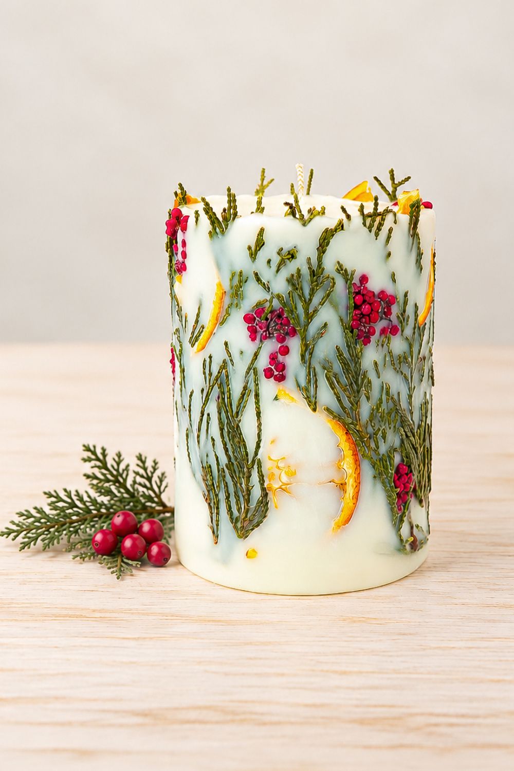 Christmas soy wax pillar candle decorated with dried orange slices, green thuja and tiny red flowers, standing on pale wood beside an evergreen sprig and red berries.