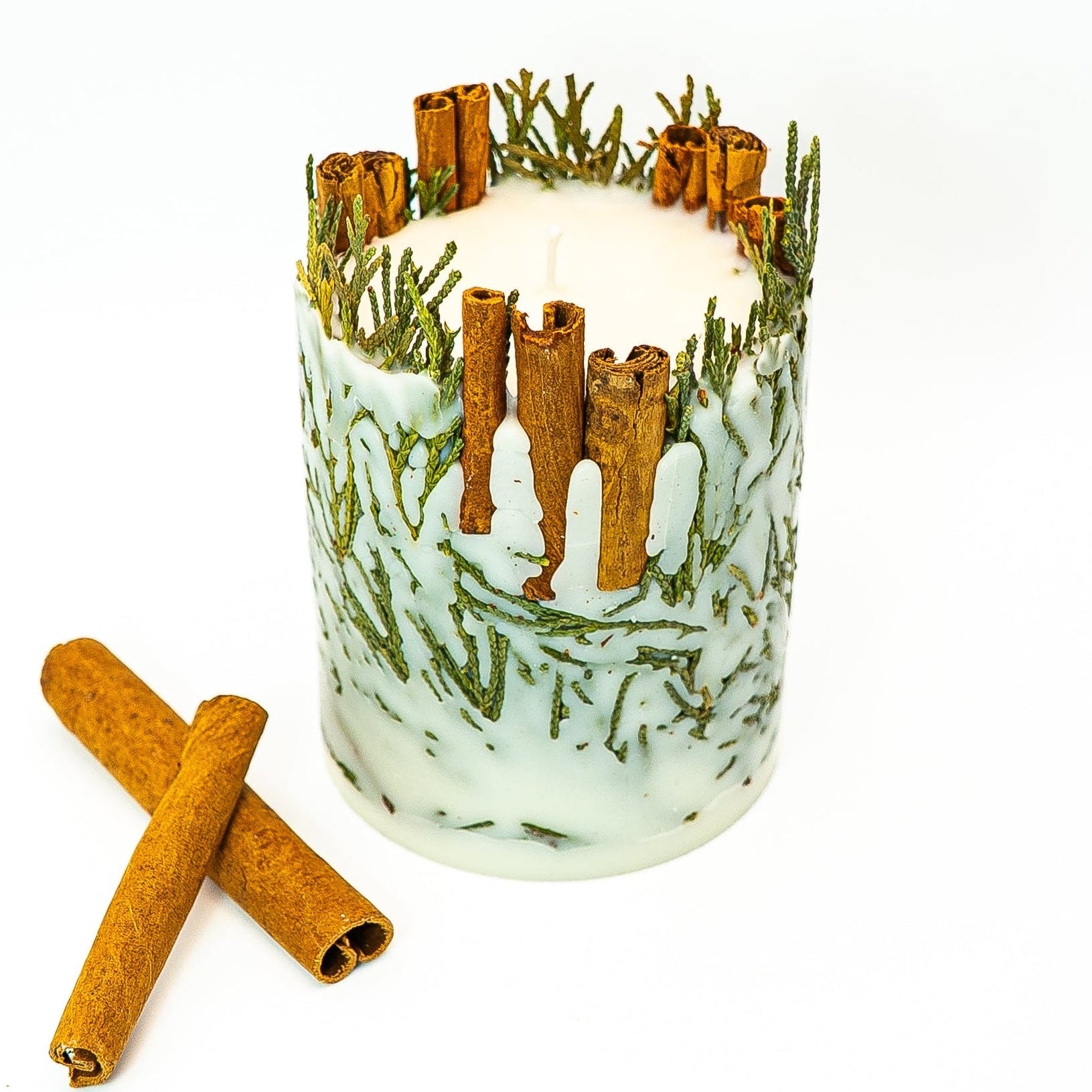Hand-poured soy wax botanical pillar candle with cinnamon sticks and evergreen sprigs embedded in wax.