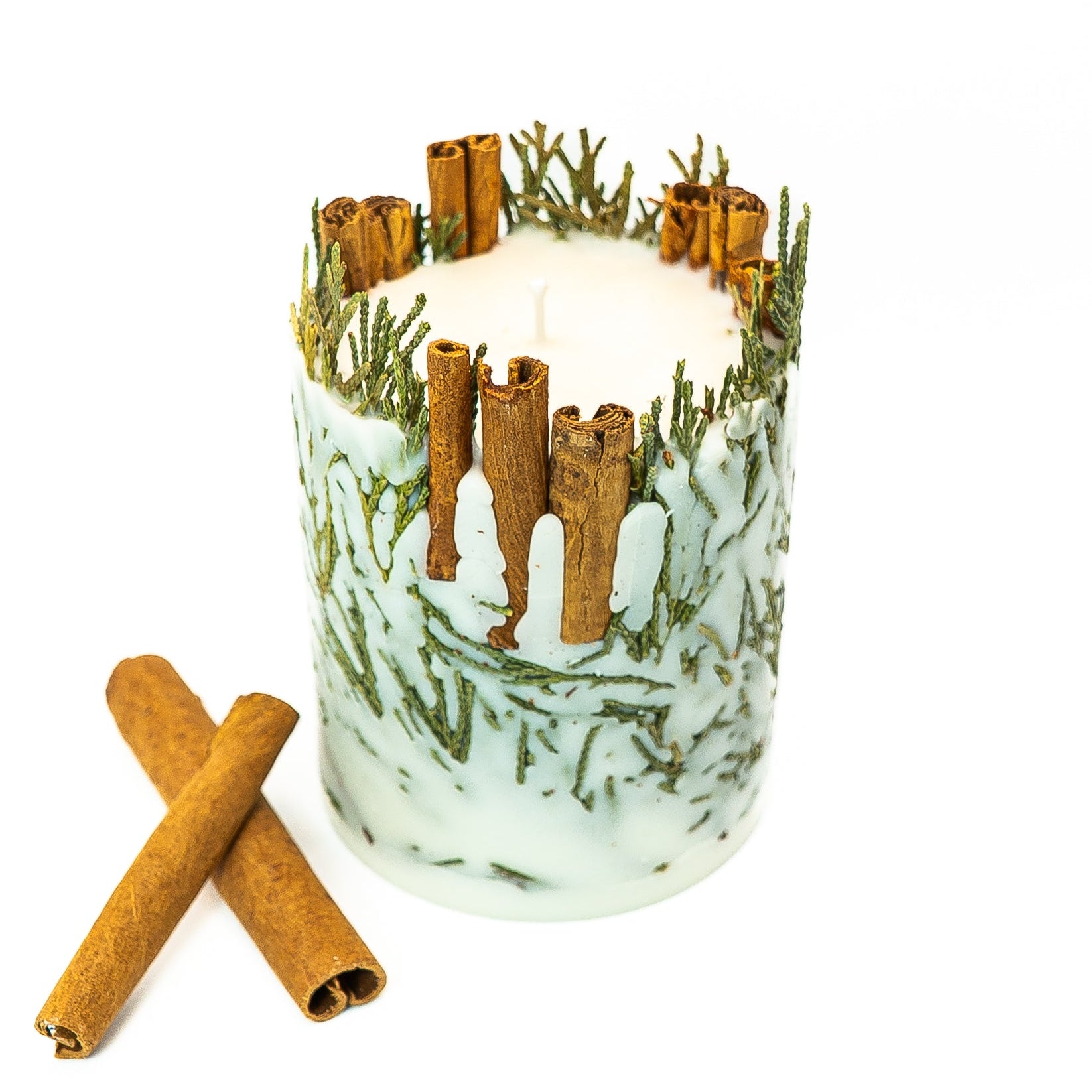 Decorative botanical pillar candle with cinnamon sticks and evergreen foliage embedded in soy wax.