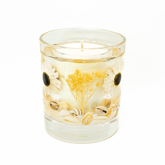 Handmade candle with soy wax center and clear gel outer layer, decorated with white daisy flowers, dried yellow flowers, and seashells in a glass jar.